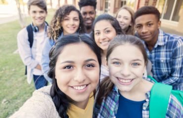 Diverse group of eight high school students are smiling and looking at the camera. Teenagers are students at public high school, and are wearing backpacks or holding school books.